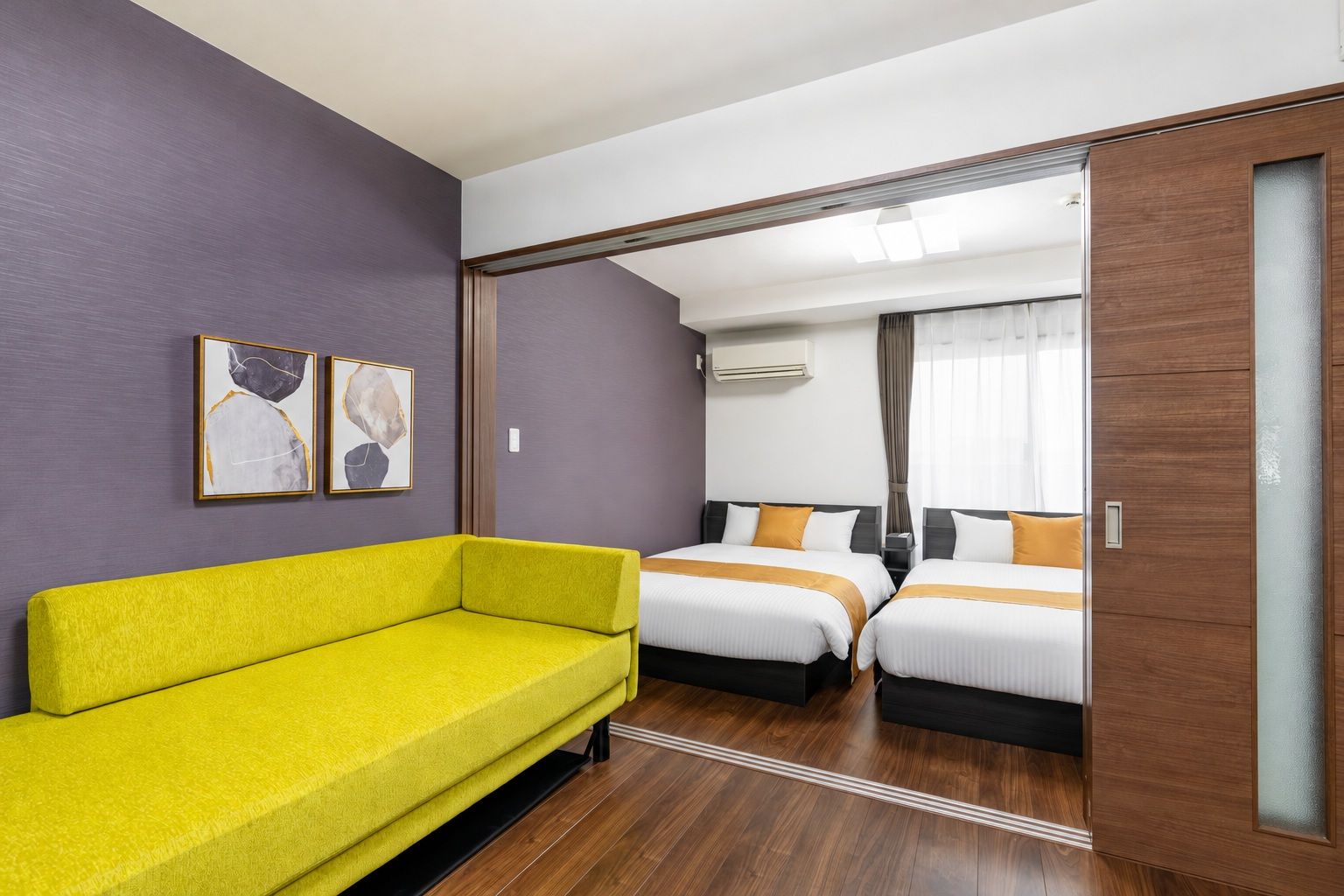 A hotel room with a yellow sofa, two beds with white and orange bedding, a wooden floor, and a wall-mounted air conditioner.