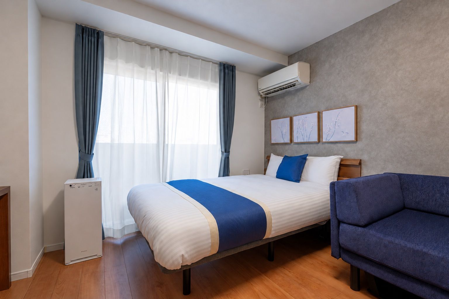 A hotel room with a bed, a blue armchair, an air conditioning unit, and a window with curtains.