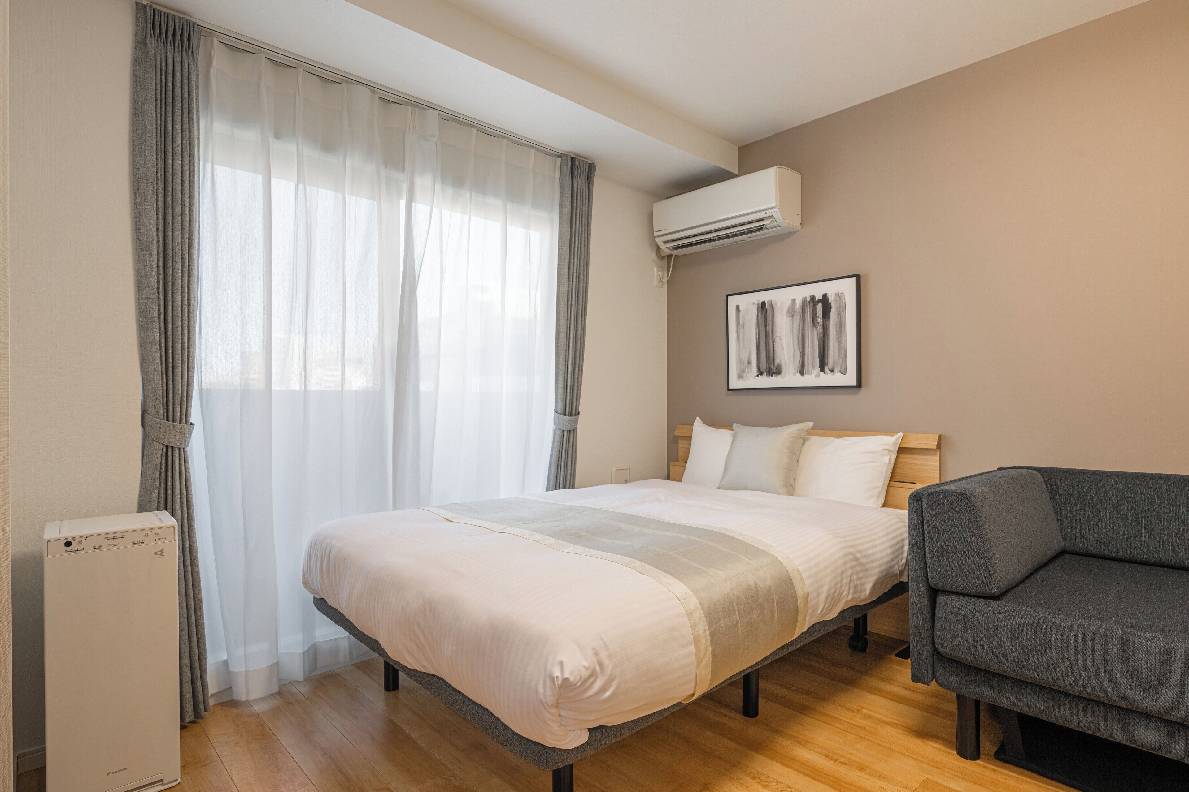 Un dormitorio con una cama, un sofá gris, un aire acondicionado blanco y una obra de arte enmarcada en la pared.