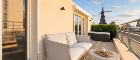 Un balcon avec un canapé blanc, une petite table ronde sur laquelle se trouvent une théière et une tasse, et un moulin à vent en arrière-plan.