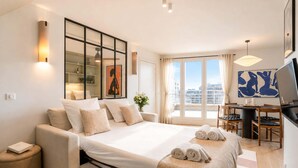 A modern hotel room with a bed, a desk, a chair, a TV, and a large window with a city view.