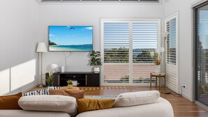 A modern living room with a large flat-screen TV, a sofa, a coffee table, and a balcony with a view of the sea. - The Family Tide – large family home, entertaining deck with views, swimming pool, air conditioning (Corlette)
