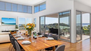 A modern dining area with a wooden table set for four, large windows with blinds, a flat-screen TV, and a view of a coastal landscape. - The Family Tide – large family home, entertaining deck with views, swimming pool, air conditioning (Corlette)