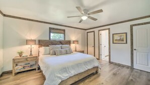 Une chambre à coucher avec un grand lit, une tête de lit en bois, des tables de chevet avec des lampes, un ventilateur de plafond et une porte ornée d’un tableau encadré.