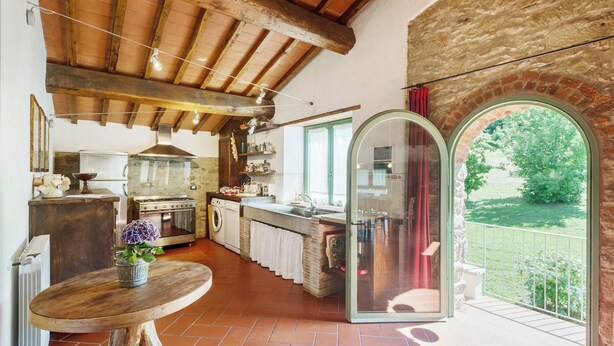 A rustic kitchen with wooden beams, a washing machine, and a large window overlooking a garden.