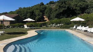 A swimming pool with white lounge chairs and umbrellas, surrounded by greenery and a stone building.