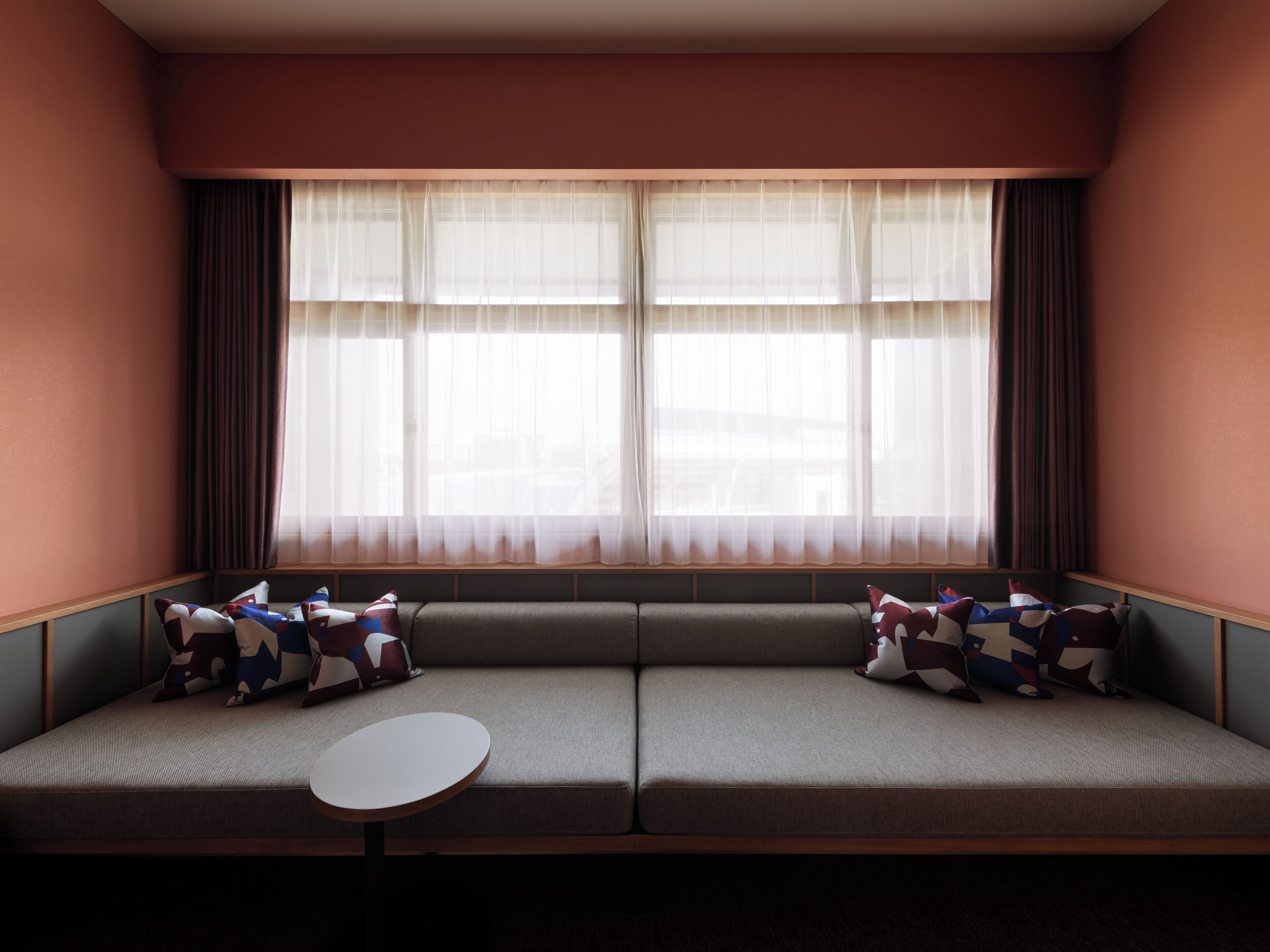 A room with a sofa facing a window with sheer curtains, a small round table in front of the sofa, and a patterned pillow on the sofa.