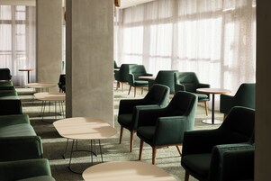 Um interior moderno com cadeiras estofadas em verde e mesas redondas, amplas janelas com cortinas leves, e um pilar de concreto.