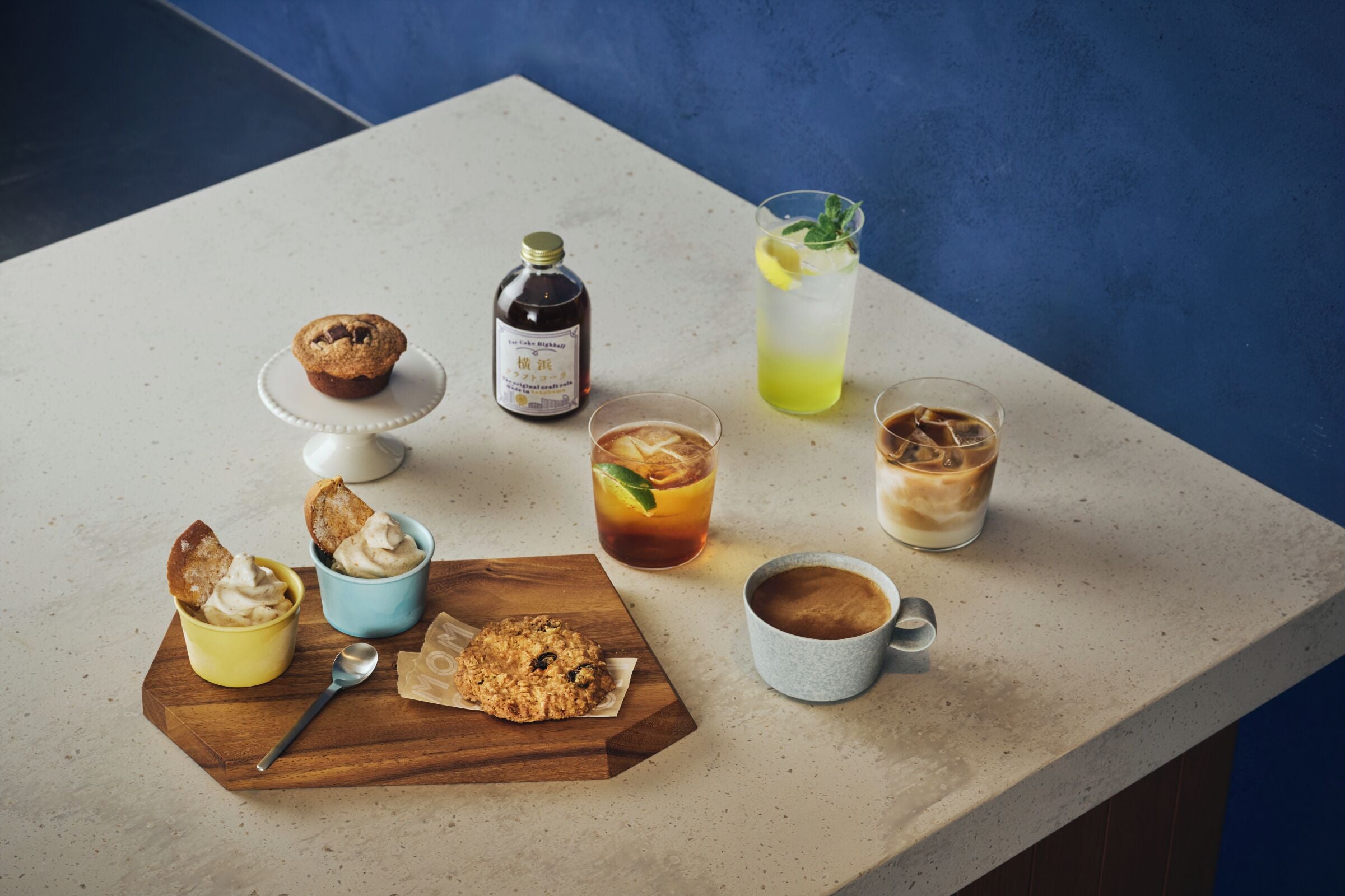 A table with a muffin, a bottle, a glass with a lime, iced drinks, a cookie, and a cup of coffee.