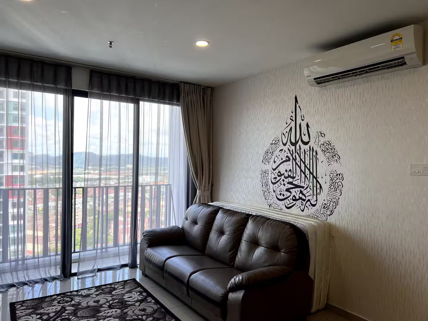 Un salon moderne avec un canapé en cuir, une grande fenêtre avec des rideaux et un mur orné de calligraphie arabe.