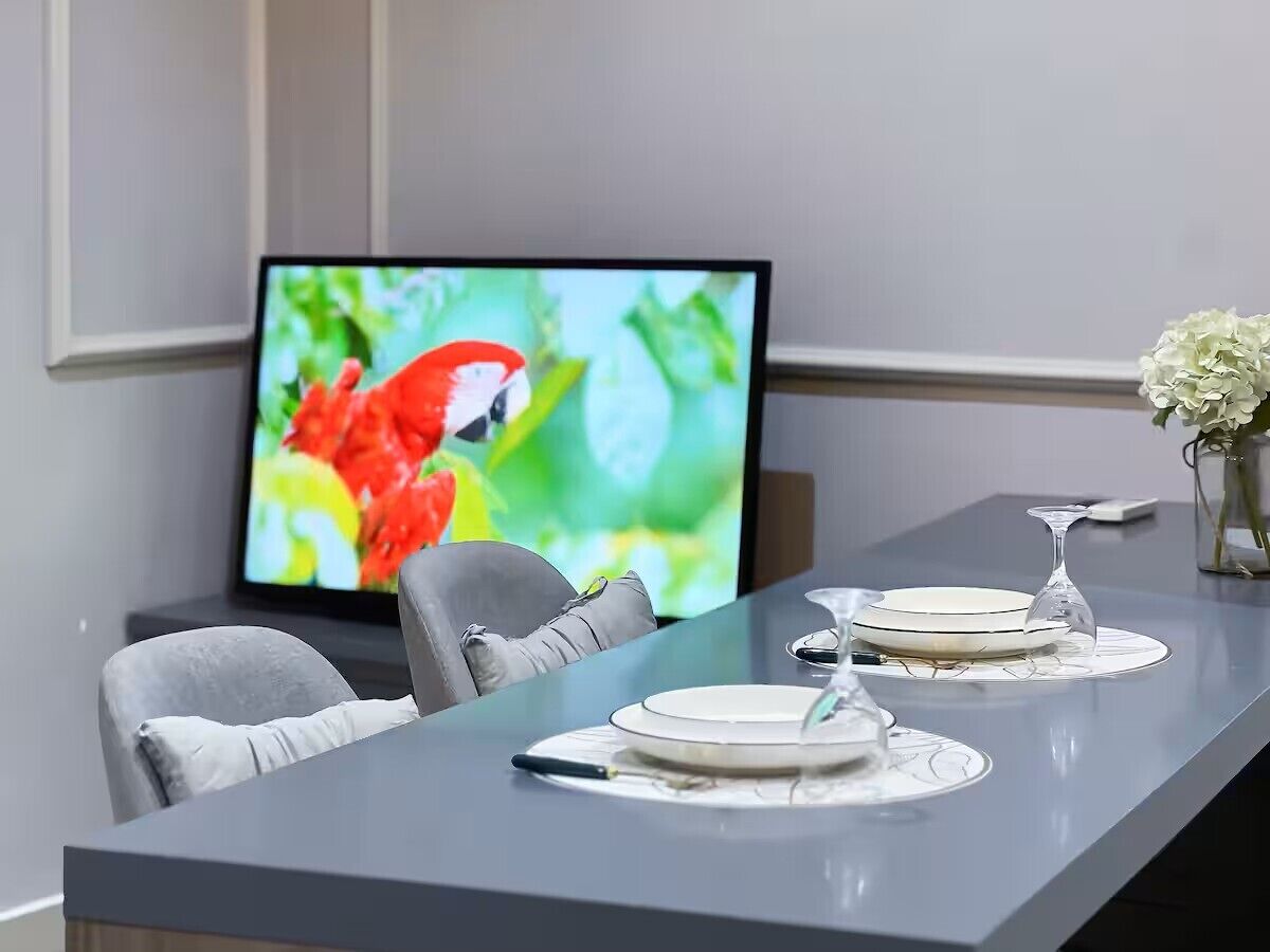 A dining area with a long table set for two, a television displaying a colorful image, and a vase with white flowers on the table.