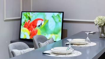 A dining area with a long table set for two, a television displaying a colorful image, and a vase with white flowers on the table.