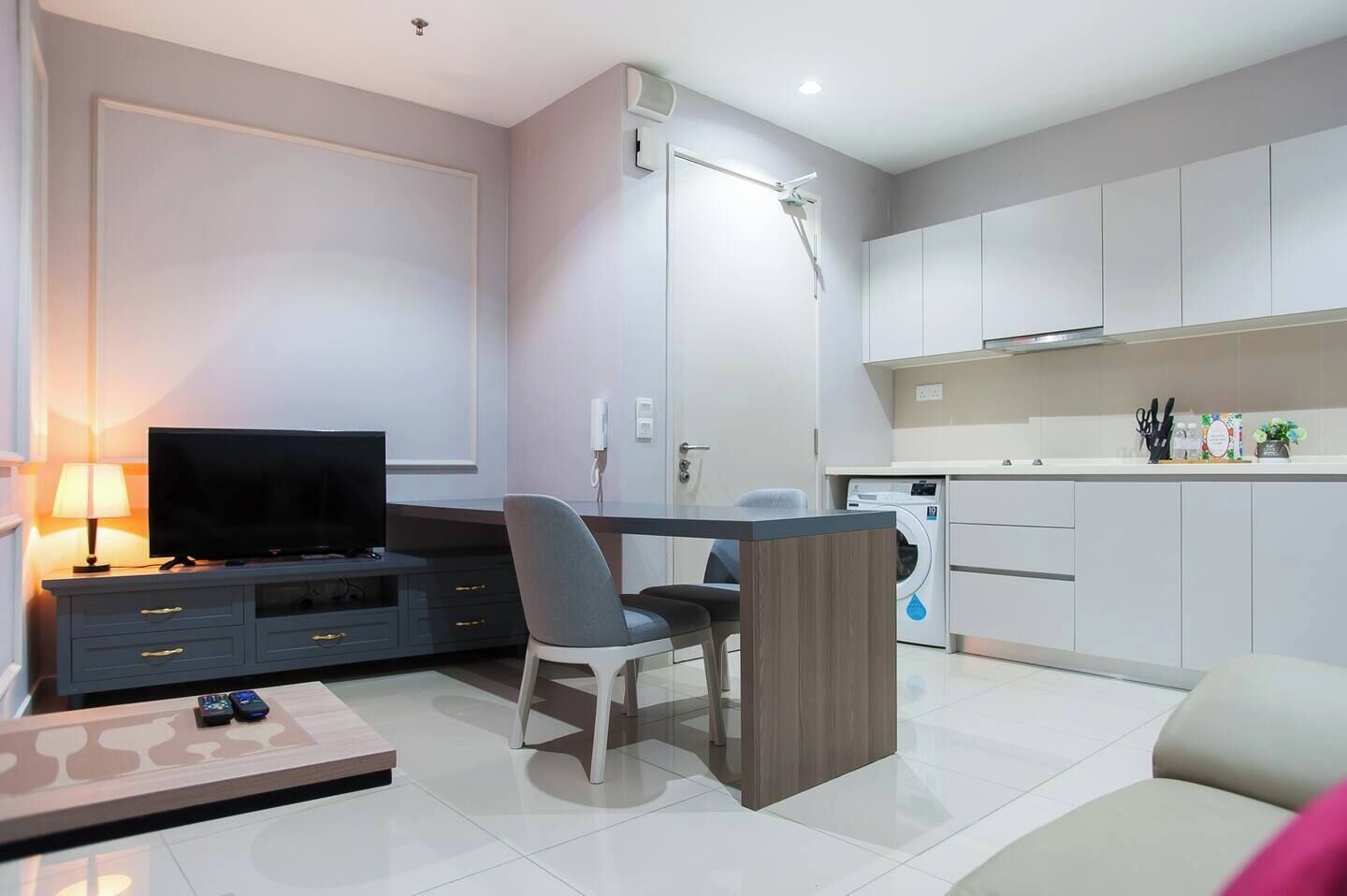 A modern living room with a flat-screen TV, a dining table with chairs, a sofa, and a coffee table. There is a washing machine in the background.
