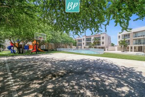 A playground with slides and a basketball hoop, surrounded by trees and modern buildings.