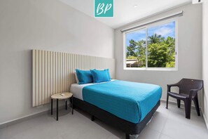 A modern bedroom with a bed, a chair, a small table, and a window with a view of trees. - 3BR Family Villa Near Beach with Pool and Parking (Bucerías)