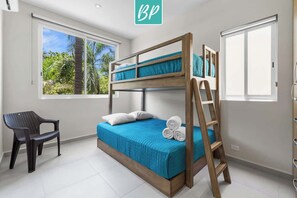 Ein Stockbett mit Holzrahmen, einer blauen Bettdecke und einer Leiter. Ein Stuhl und ein Fenster mit Blick auf Grün sind ebenfalls vorhanden.