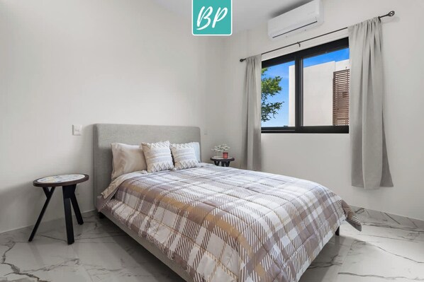 A bedroom with a bed, a small table, a window with curtains, and an air conditioning unit. - 2BR w/ Pool, Fast WiFi & Beach Gear - Stanza 7 (Bucerías)