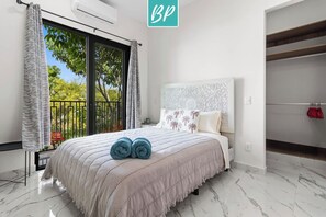 A bedroom with a large bed, a window with curtains, a white headboard, and a view of trees outside.