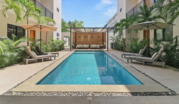 A swimming pool with lounge chairs and umbrellas, surrounded by greenery and a building with balconies. - 2BR w/ Pool, Fast WiFi & Beach Gear - Stanza 7 (Bucerías)