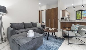 Una sala moderna con un sofá gris, una mesa de centro redonda y un sillón gris. Se observa un área de cocina con ventana y lámparas colgantes.