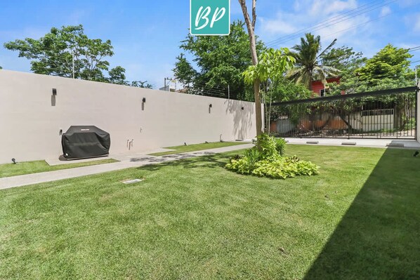 Un área exterior bien cuidada con un camino pavimentado, una parrilla cubierta y un pequeño jardín con una palmera.
