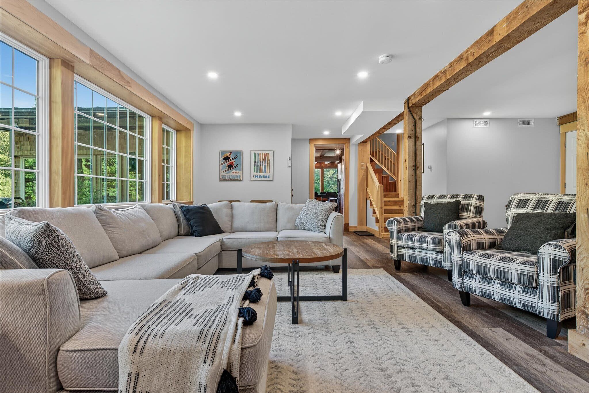 A spacious living room with a large sectional sofa, a round coffee table, and patterned armchairs. There are large windows with wooden frames, a staircase leading to another level, and artwork on the walls.