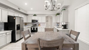 A modern kitchen with white cabinetry, stainless steel appliances, a wooden dining table, and a chandelier.