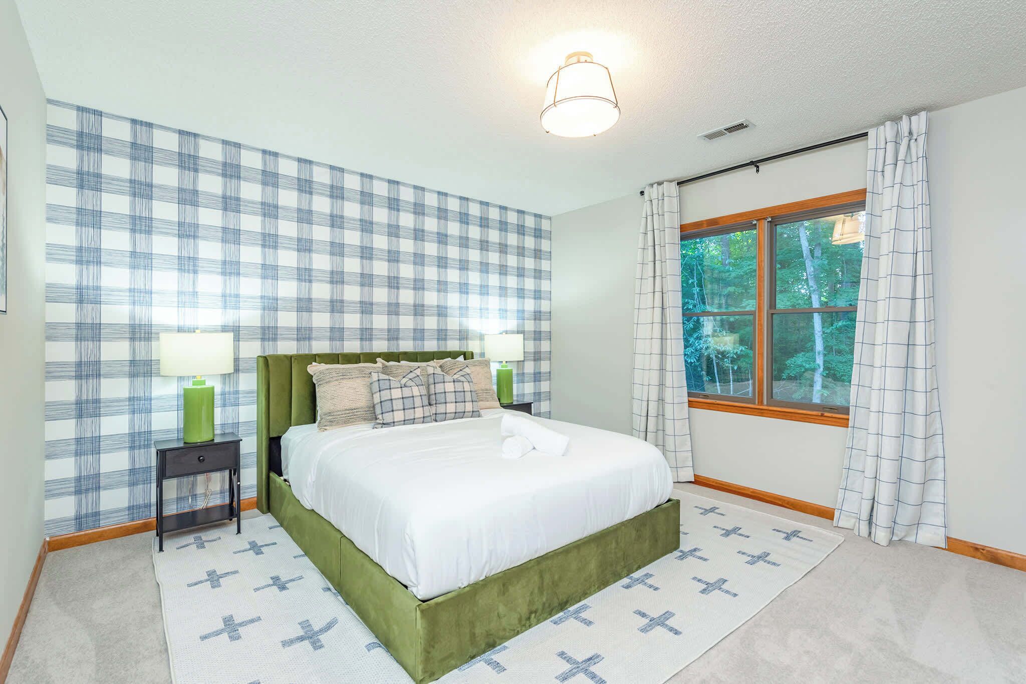 A bedroom with a green bed frame, a white mattress, a nightstand with a lamp, a window with curtains, and a checkered wall.