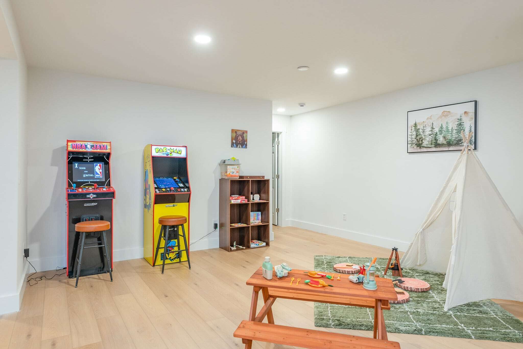 A room with two arcade machines, a small bookshelf, a wooden table with toys, and a tent-like structure.