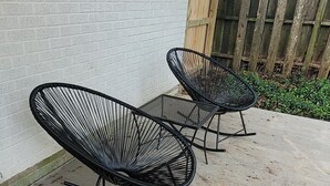 Two black rocking chairs with metal frames and mesh backs, positioned on a concrete patio next to a white brick wall and a wooden fence. - Mid City Modern, Good Vibes in BRLA (Baton Rouge)