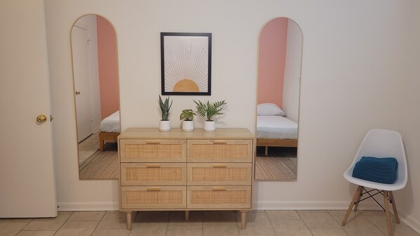 A room with a wooden dresser, two beds, a chair, and a framed artwork on the wall.