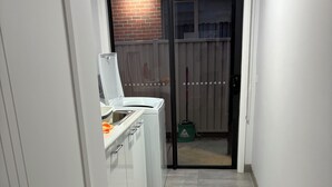 A narrow hallway with a kitchen area featuring a white washing machine and a sink. A sliding glass door leads to an outdoor area with a brick wall and a fence. - Country Retreat (Beaufort)