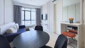 Ein modernes Hotelzimmer mit einer Couch, einem Esstisch mit Stühlen, einem an der Wand befestigten Fernseher und einem großen Fenster mit Vorhängen.
