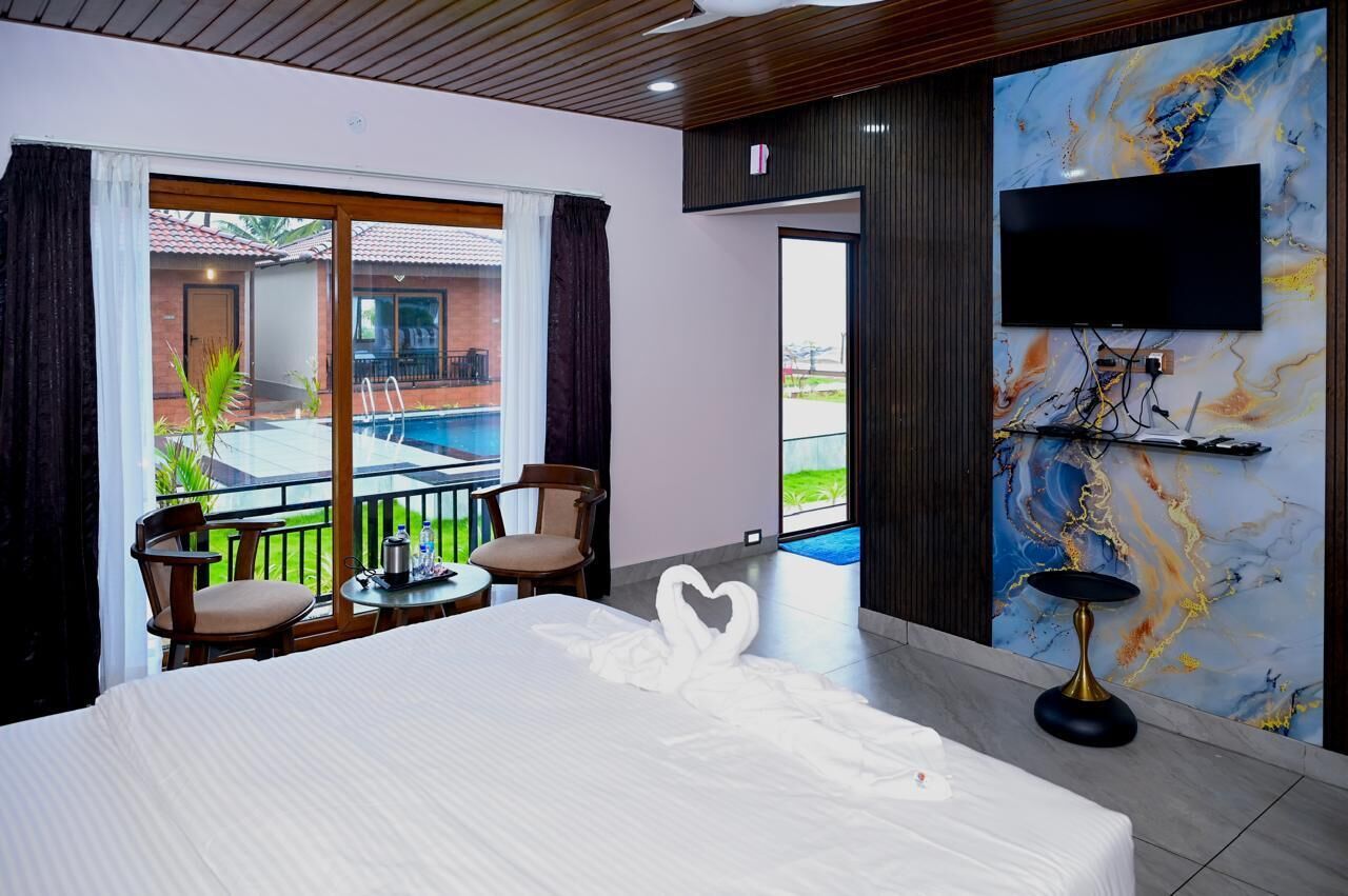 A hotel room with a large bed, a TV mounted on a marble wall, a balcony with a pool, and a view of a building with a red roof.