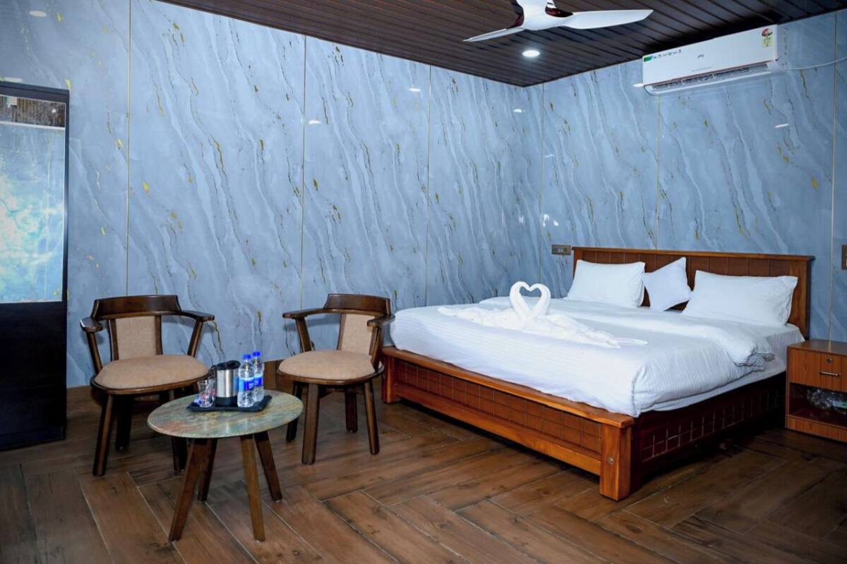 A hotel room with a wooden bed, two chairs, a small round table with a bottle and glass, and a ceiling fan.
