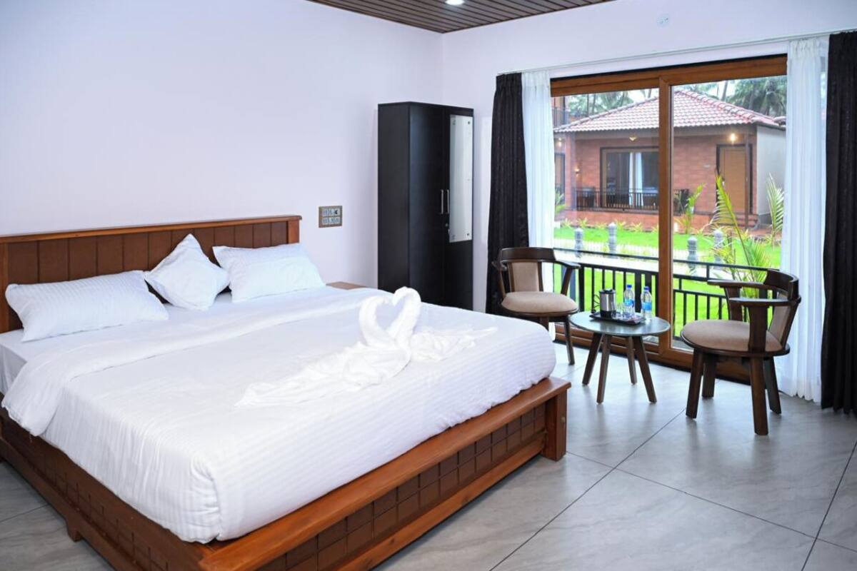 A hotel room with a large bed, a TV, a balcony with chairs, and a view of a building outside.
