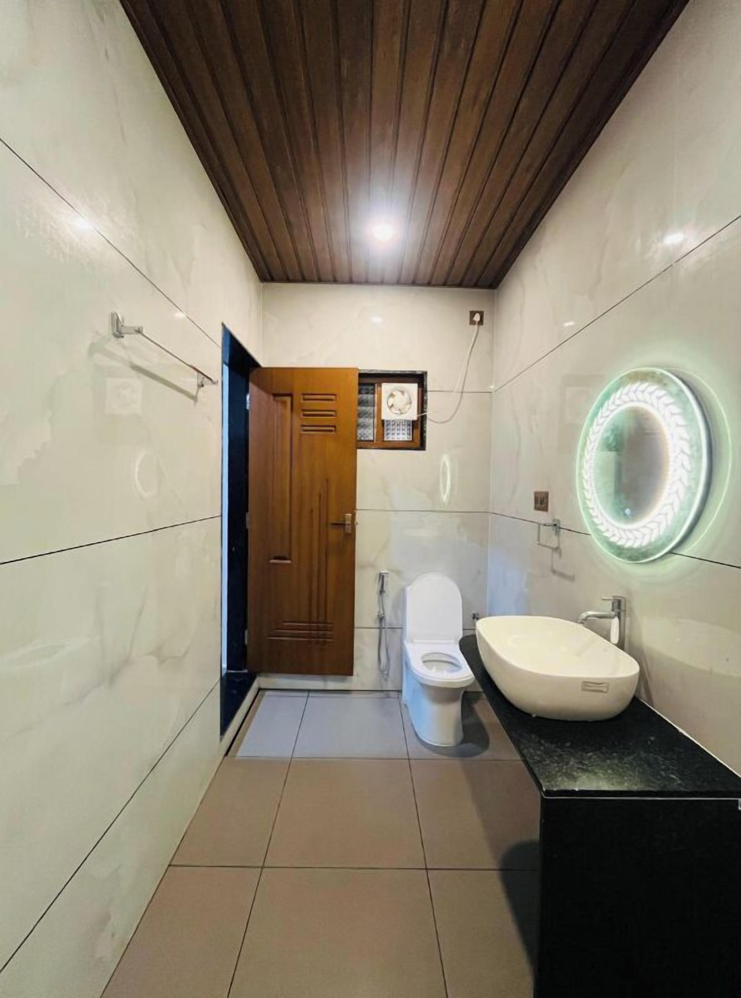 A bathroom with a wooden door, a toilet, a sink with a circular mirror, and a tiled floor.