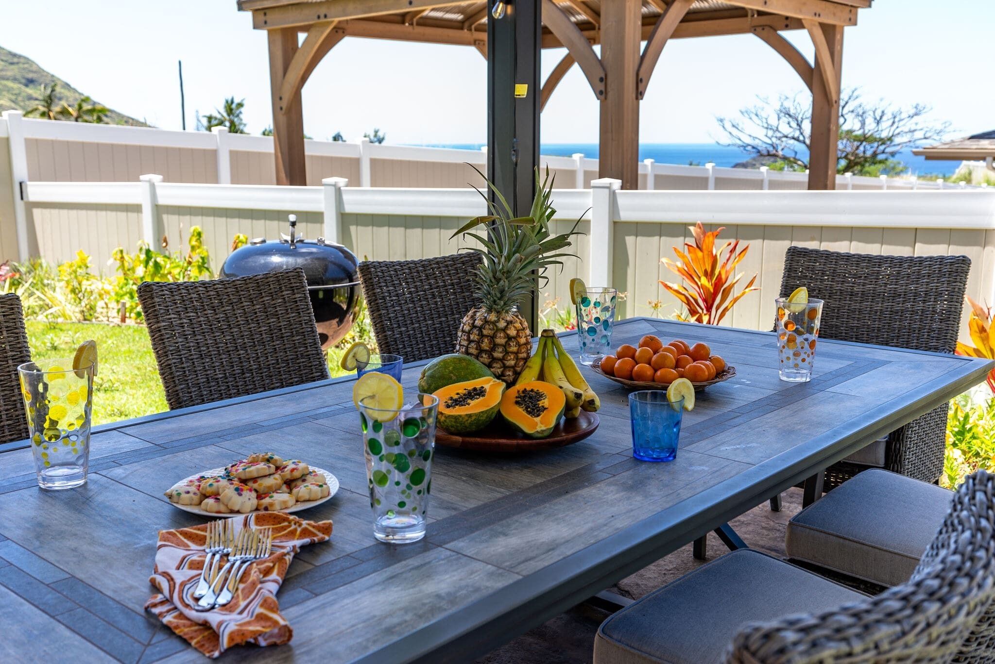Un espace extérieur aménagé pour les repas, avec des chaises en osier, une table dressée avec des fruits, des boissons et un barbecue.