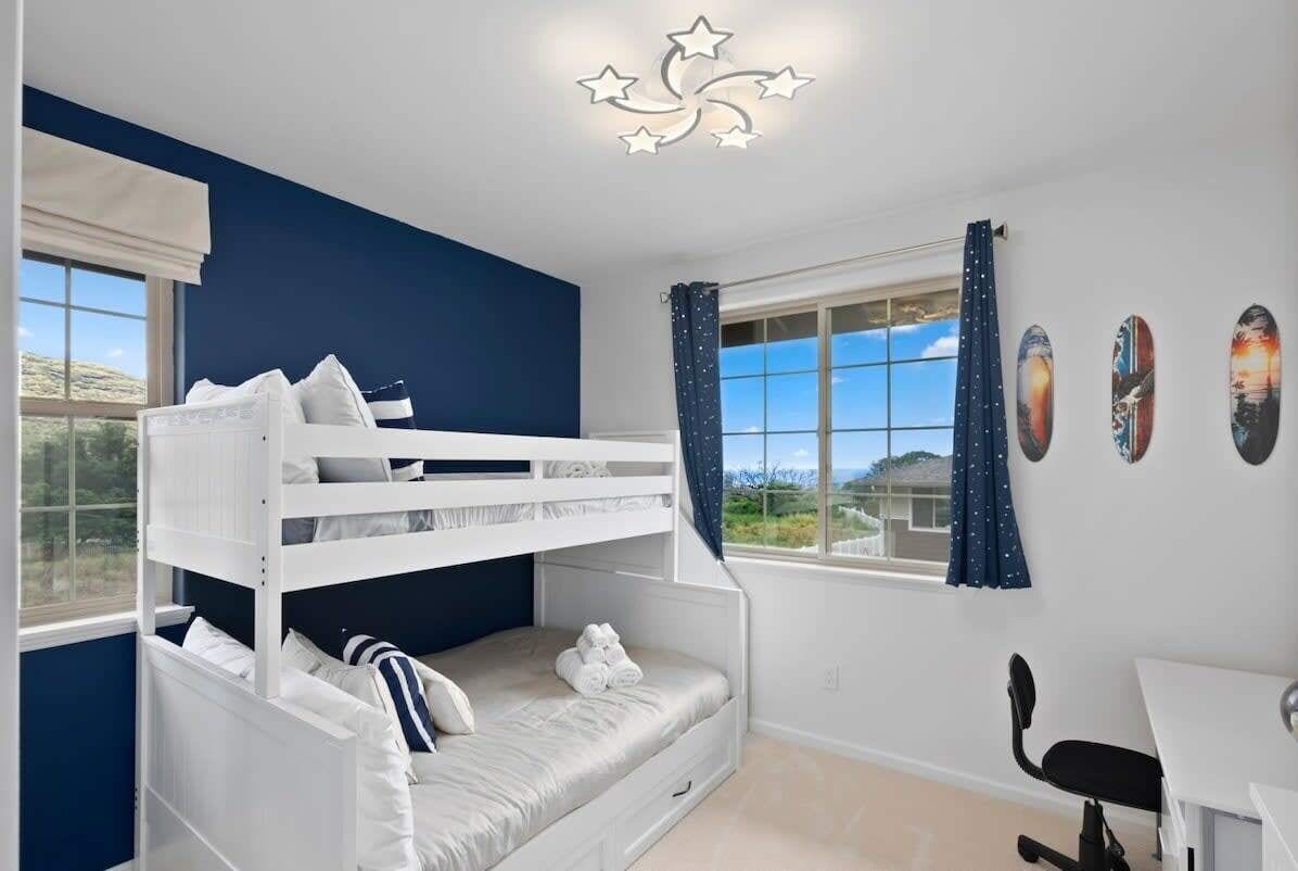 A room with a bunk bed, a desk, and a chair. There are windows with curtains, and a ceiling light fixture.