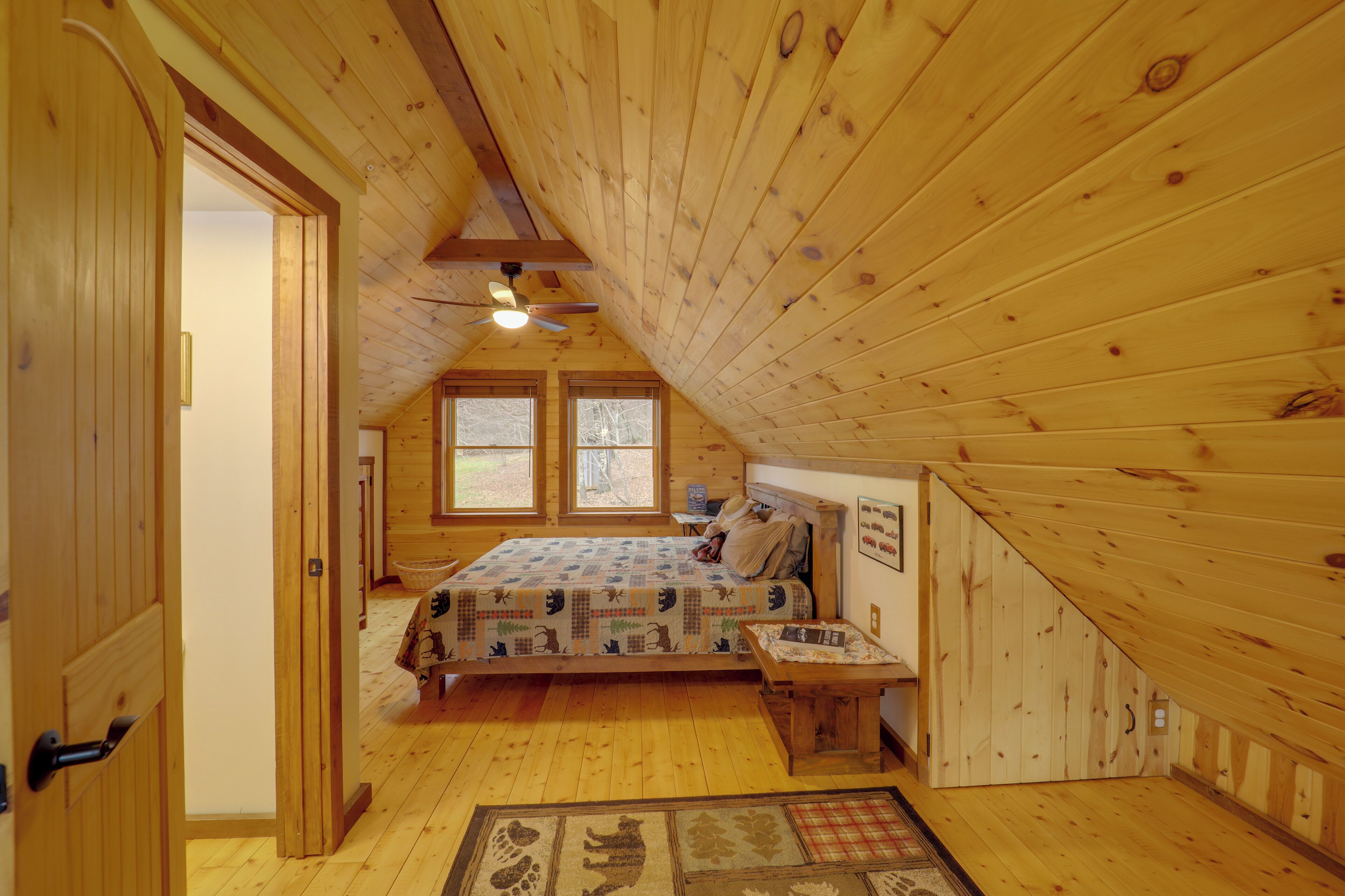 A cozy attic bedroom with a wooden ceiling, a bed with a patterned bedspread, a small table, and a window with a view of trees.