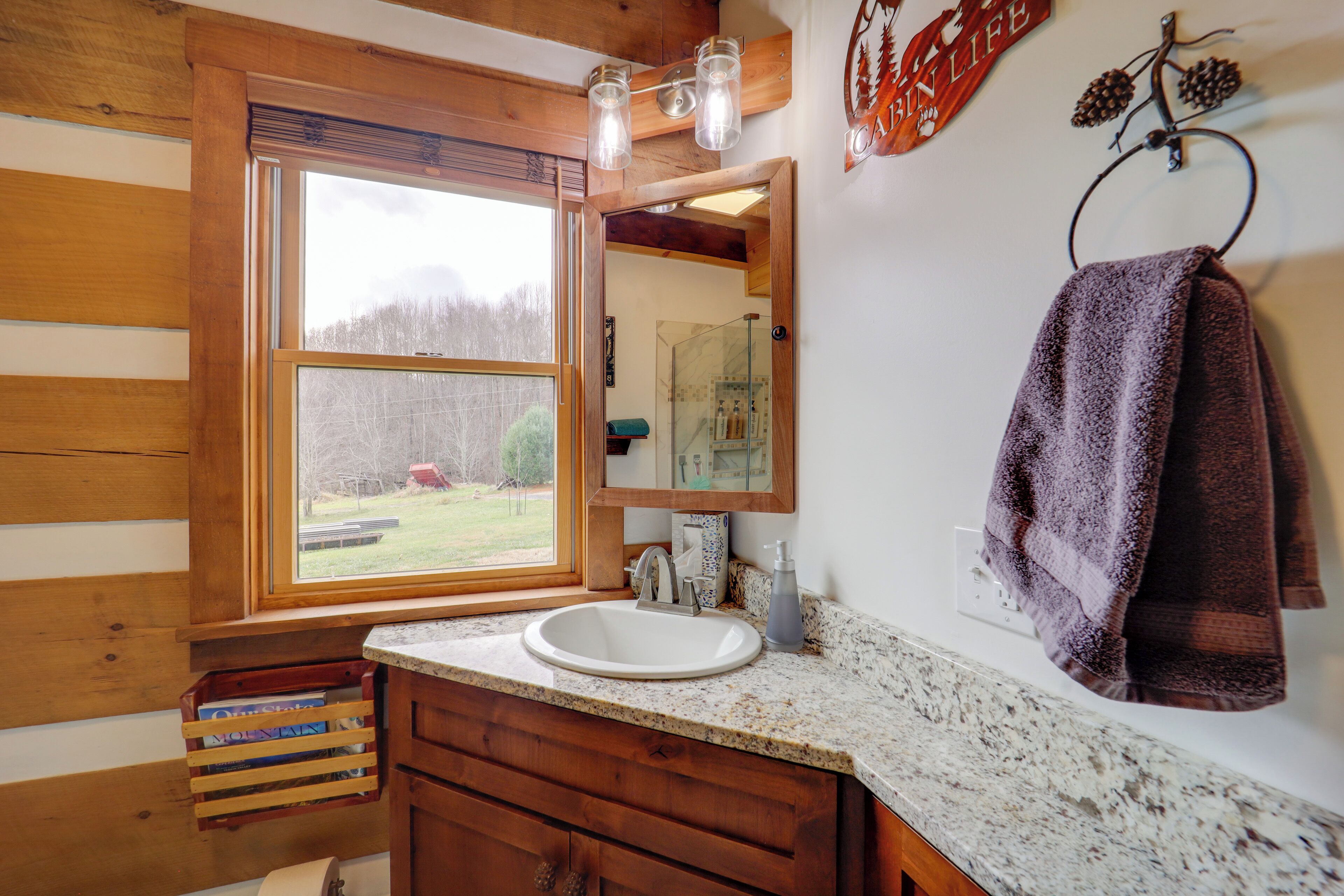 A wooden cabin bathroom with a large window, a white sink, wooden cabinets, and a towel rack.