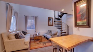 A living room with a sofa, coffee table, and a wooden dining table. A spiral staircase is visible in the background.