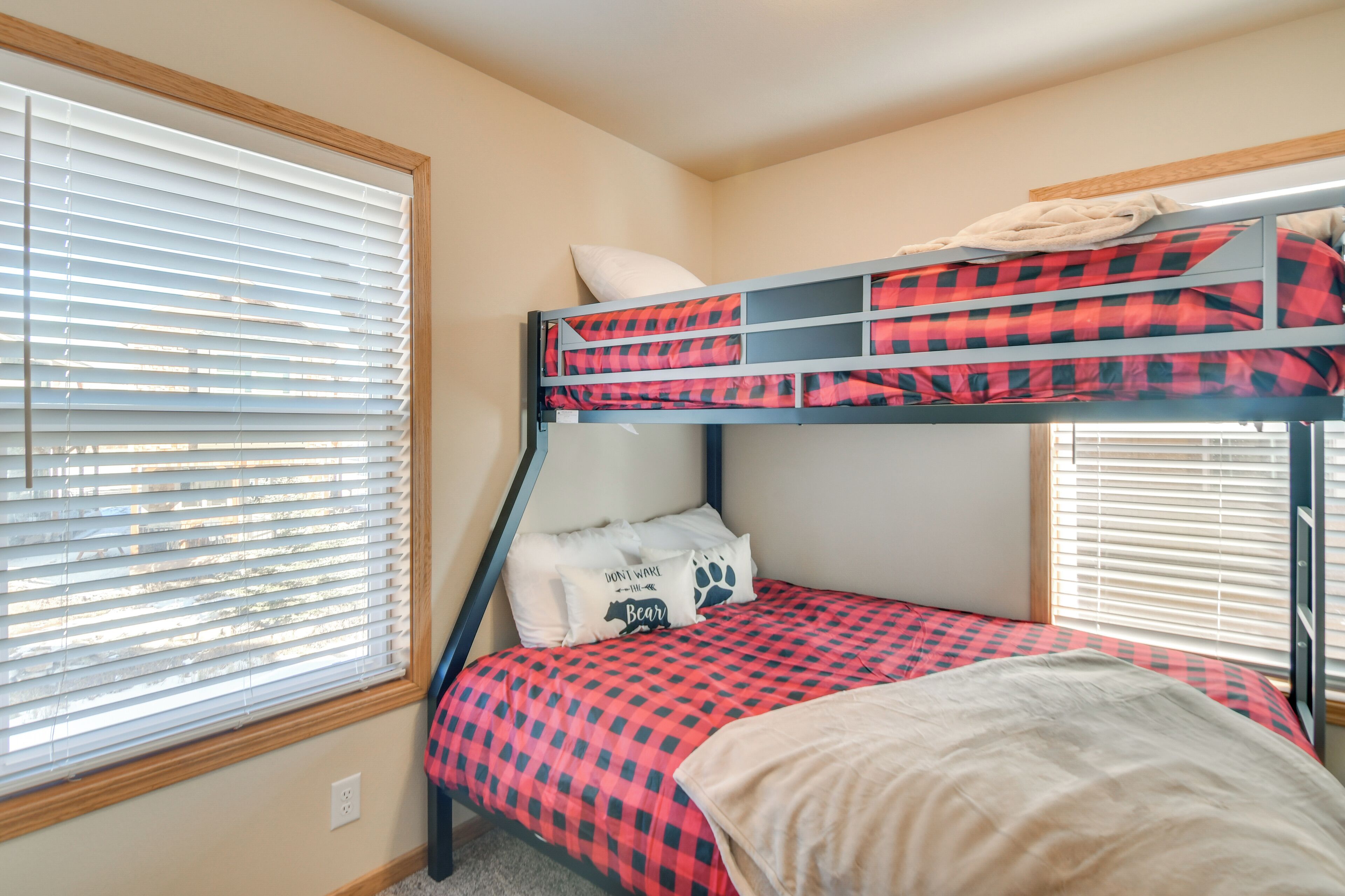 A bunk bed room with a checkered bedspread, a pillow with a bear design, and a window with blinds.