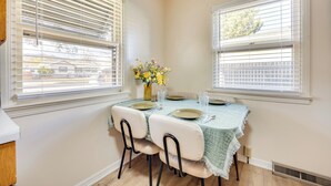 Un coin repas avec une table dressée pour quatre personnes, deux fenêtres équipées de stores, un vase avec des fleurs et une vue sur les maisons à l’extérieur.
