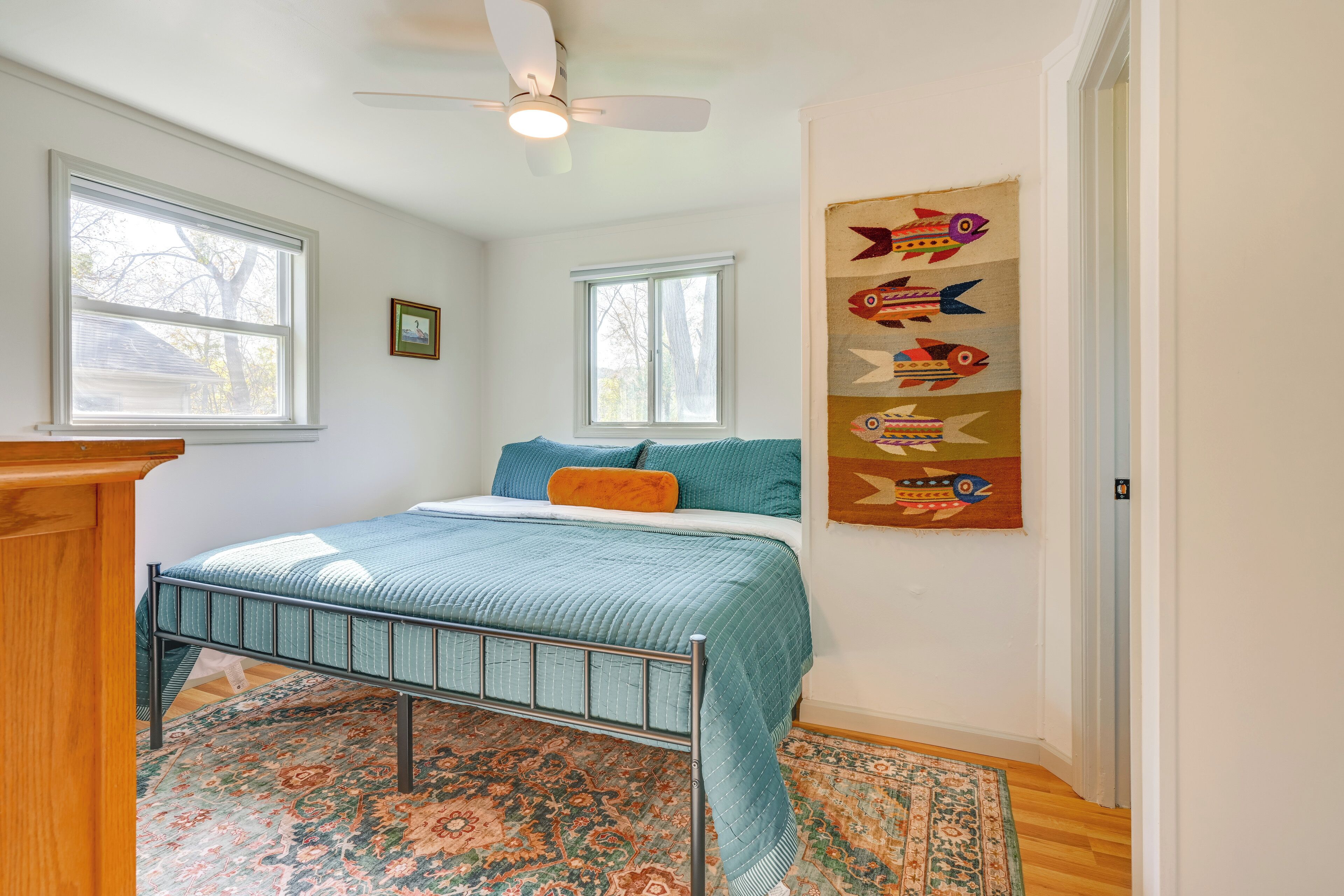 A bedroom with a bed, two windows, a ceiling fan, and a wall hanging.