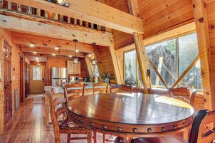 Un interior de madera con una mesa redonda rodeada de sillas, una zona de cocina y ventanas amplias.