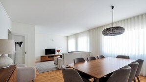A modern living room with a wooden dining table, grey upholstered chairs, a sofa, a TV, and a large window with sheer curtains. - Acm021 Bombarda Homes, Spacious 3BR for Families (Porto)