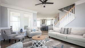 A spacious living room with a high ceiling fan, a staircase, and a view to the kitchen.