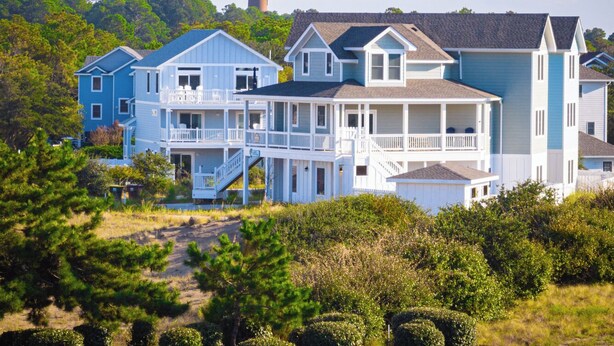 A coastal house with multiple levels, a prominent lighthouse in the background, and a beach in the foreground.