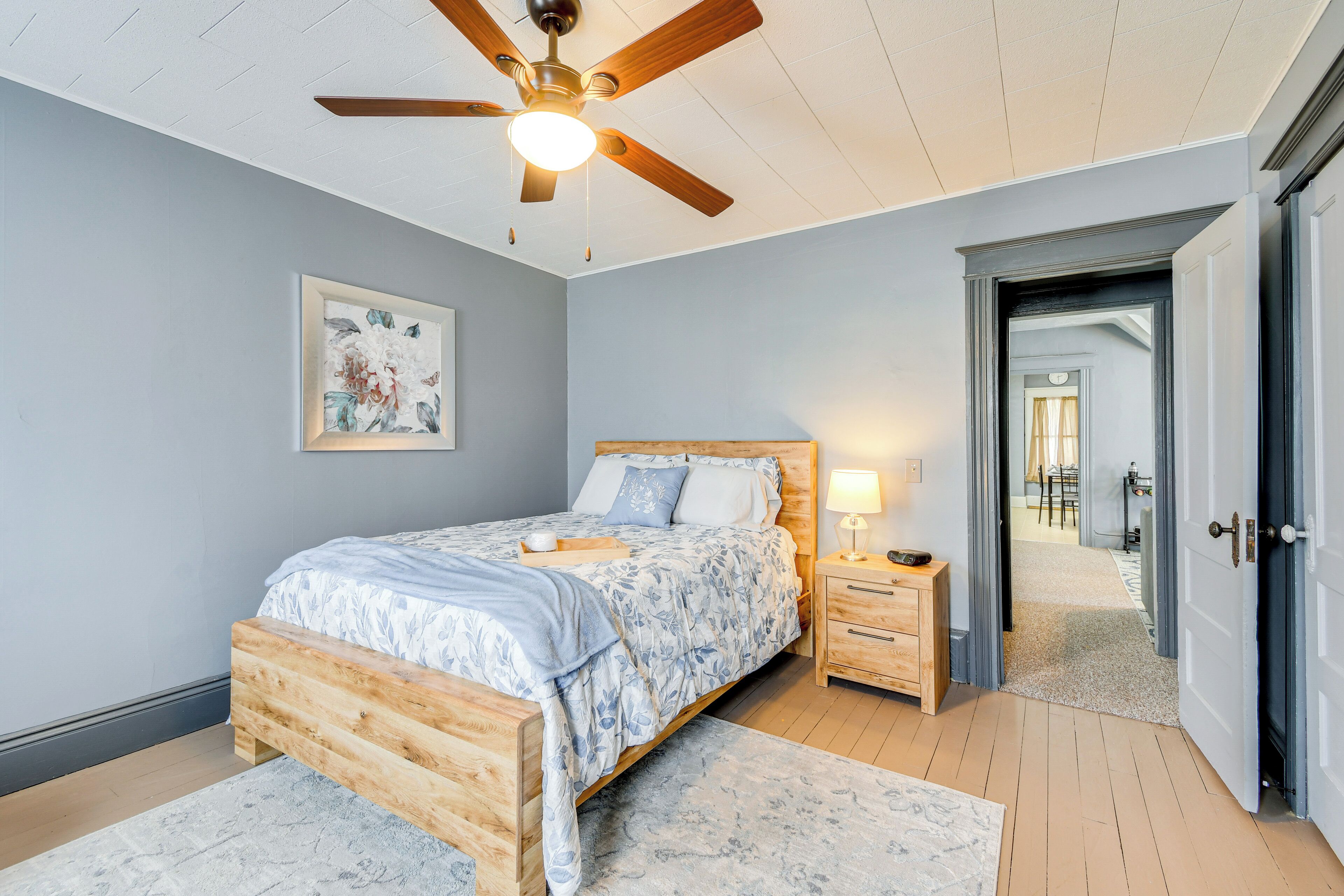 A bedroom with a bed, wooden nightstands, a ceiling fan, and a door leading to another room.